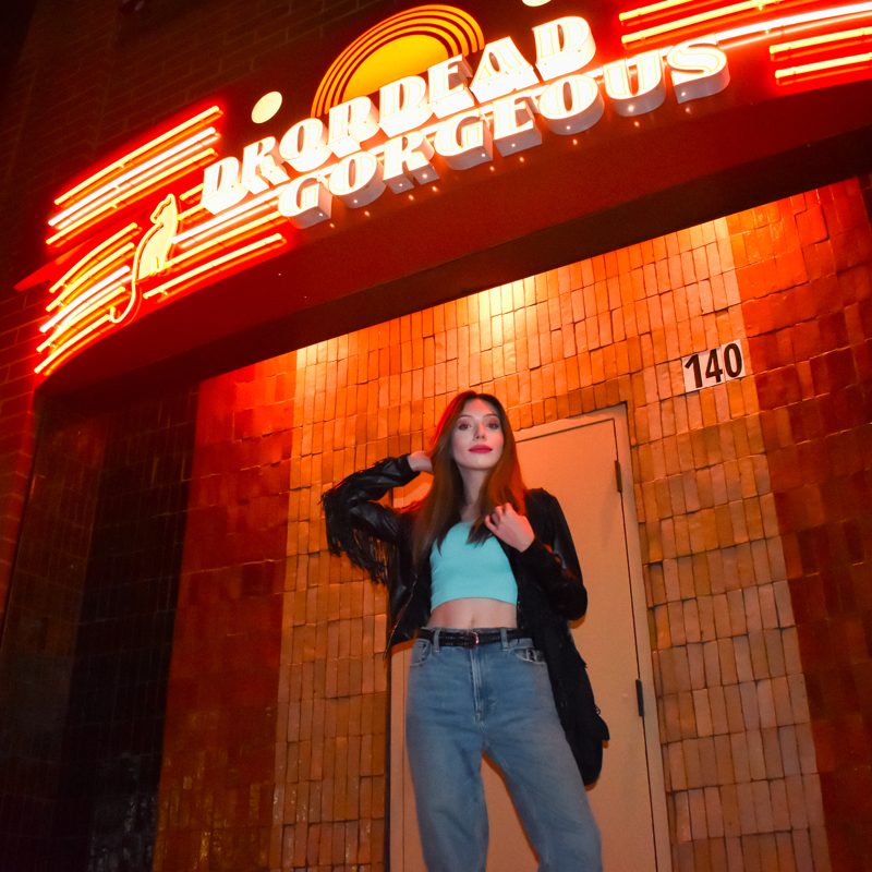 Rose standing under a neon sign that says Drop Dead Gorgeous